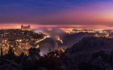 Desde arriba asombroso paisaje del antiguo castillo iluminado del Alcázar de Toledo sobre la ciudad en el crepúsculo brumoso - foto de stock