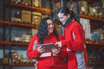 D'en bas de jeunes femmes en uniforme rouge souriant tout en utilisant une tablette numérique pendant le travail dans un entrepôt moderne — Photo de stock