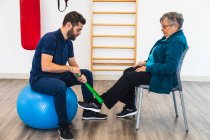 Side view of male personal coach sitting on exercise blue ball while using green elastic band on ankle of old woman in gym — Stock Photo