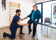 Young bearded male personal trainer kneeling while holding hand and knee of aged woman during exercising with cane in gym — Stock Photo