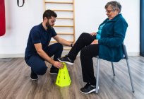 Side view of aged woman in sportswear sitting on chair while putting foot on yellow fitness cone with personal male trainer in gym — Stock Photo
