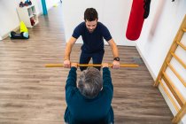 Visão traseira de cima de senhora sênior segurando vara com treinador pessoal masculino durante o treino de fisioterapia no ginásio — Fotografia de Stock