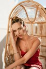 Sensuale bionda dai capelli bella modello femminile con orecchini d'oro in abito rosso elegante seduto sulla sedia appesa sulla terrazza all'aperto alla luce del sole — Foto stock
