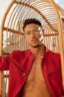 Serious handsome man with stylish hairdo in trendy eyeglasses and red jacket sitting on hanging chair and looking at camera — Stock Photo