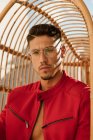Serious handsome man with stylish hairdo in trendy eyeglasses and red jacket sitting on hanging chair and looking at camera — Stock Photo