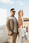 Jeune homme en costume tendance et lunettes de soleil et femme en pantalon et soutien-gorge debout près de la lumière du soleil — Photo de stock