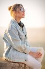 Vista laterale di giovane donna in abbigliamento casual con occhiali da sole poggiati su recinzione rocciosa con bevanda e musica d'ascolto con cuffie — Foto stock