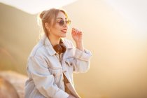 Радісна молода жінка з сонцезахисними окулярами в модному повсякденному вбранні посміхається і дивиться в сонячний день — стокове фото