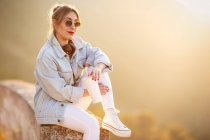 Удовлетворенная блондинка в модных солнцезащитных очках и повседневной одежде сидит на каменистом заборе и смотрит на камеру в солнечном свете на размытом фоне — стоковое фото