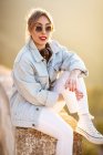 Satisfait dame blonde aux lunettes de soleil à la mode et vêtements décontractés assis sur une clôture rocheuse et regardant la caméra en plein soleil sur fond flou — Photo de stock