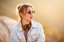 Fröhliche junge Frau mit Sonnenbrille im trendigen lässigen Outfit lächelt und schaut an sonnigen Tagen weg — Stockfoto