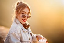 Радостная молодая женщина в солнечных очках в модном повседневном наряде улыбается и смотрит в камеру в солнечный день — стоковое фото