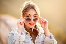 Joyful young woman with sunglasses in trendy casual outfit smiling and looking at camera on sunny day — Stock Photo
