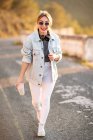 Joyful blond haired woman in stylish outfit and sunglasses walking with beverage and smiling at camera on blurred background — Stock Photo