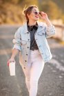 Mulher loira alegre de cabelos loiros em roupa elegante e óculos de sol andando com bebida e sorrindo no fundo borrado — Fotografia de Stock