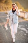 Mulher loira alegre de cabelos ruivos em roupa elegante e óculos de sol andando com bebida e sorrindo para a câmera no fundo borrado — Fotografia de Stock