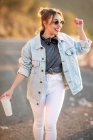 Joyful blond haired woman in stylish outfit and sunglasses walking with beverage and smiling on blurred background — Stock Photo