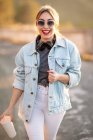 Joyful blond haired woman in stylish outfit and sunglasses walking with beverage and smiling on blurred background — Stock Photo