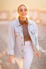Mulher loira alegre de cabelos loiros em roupa elegante e óculos de sol andando com bebida e sorrindo no fundo borrado — Fotografia de Stock