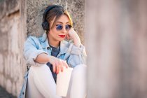 Jeune femme en tenue décontractée avec des lunettes de soleil reposant sur une clôture rocheuse avec des boissons et écouter de la musique avec des écouteurs — Photo de stock