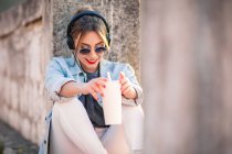 Junge, fröhliche Frau in Freizeitkleidung mit Sonnenbrille, die sich mit Getränken auf felsigen Zaun stützt und mit Kopfhörern Musik hört — Stockfoto