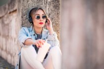 Молода жінка в повсякденному одязі з сонцезахисними окулярами відпочиває на скелястому паркані з напоєм і слухає музику з навушниками — стокове фото