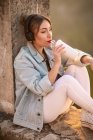 Вид сбоку на молодую женщину в повседневной одежде, отдыхающую на скалистом заборе с напитком и слушающую музыку с наушниками — стоковое фото