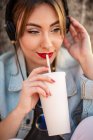 Молода жінка в повсякденному одязі відпочиває на кам'янистому паркані з напоєм і слухає музику з навушниками — стокове фото