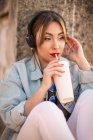 Jovem mulher em desgaste casual descansando em cerca rochosa com bebida e ouvir música com fones de ouvido — Fotografia de Stock