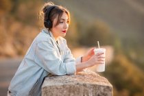Side view of young lady in trendy jeans jacket with red lips leaning elbows on fence and admiring view with headphones and beverage — Stock Photo