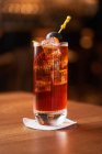 Verre Highball avec cocktail d'alcool rouge avec glaçons décorés avec un bâton d'olive noire sur un comptoir en bois — Photo de stock