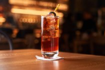 Bicchiere Highball con cocktail di alcool rosso con cubetti di ghiaccio decorato con bastone con oliva nera sul bancone di legno — Foto stock