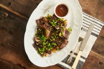 Ansicht von oben serviert leckeres gebackenes Fleisch mit grünen Zwiebeln und BBQ-Sauce auf Teller mit Besteck auf rustikalem Holztisch — Stockfoto