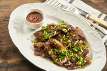 Ansicht von oben serviert leckeres gebackenes Fleisch mit grünen Zwiebeln und BBQ-Sauce auf Teller mit Besteck auf rustikalem Holztisch — Stockfoto