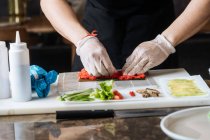Cultiver cuisinier qualifié dans des gants jetables pétrissant farce épicée rouge sur la table avec du poisson vert et des sauces dans la cuisine — Photo de stock