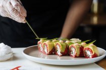 Crop cuoco in guanti usa e getta decorare sushi fresco gustoso con verdure su piatto bianco sul tavolo — Foto stock