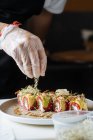 Cosechadora en guantes desechables decorando sushi fresco y sabroso con verduras en plato blanco en la mesa - foto de stock