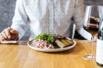 Crop person tagliare succose costole di manzo appetitoso con una forchetta e coltello in piatto con guarnire verde a tavola e bere vino nel ristorante — Foto stock
