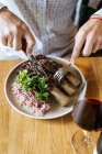 Pessoa de colheita cortando costelas de carne apetitosas suculentas com um garfo e faca em prato com guarnição verde na mesa e bebendo vinho no restaurante — Fotografia de Stock