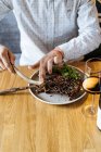 Crop person cutting juicy appetizing beef ribs with a fork and knife in plate with green garnish at table and drinking wine in restaurant — Stock Photo