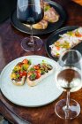 Сверху празднично украшенные красочные бутерброды с красно-желтыми нарезанными помидорами черри и травами на белой тарелке с вином на столе — стоковое фото