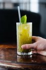 Hand o beschnitten unkenntliche Person trinkt leckere appetitlich gekühlte Limonade mit Minze und Stroh auf dem Tisch im Sonnenlicht — Stockfoto