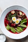 Сверху вкусный красный свекольный суп с вареными яйцами и травами в белой тарелке на столе — стоковое фото
