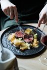 Crop from above of person with black plate with roast beef slices on creamy sauce with cauliflower and fried chips having dinner with glass of red wine — Stock Photo