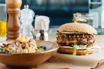 Big hamburger on paper with cutlet cheese and vegetables served with bowl of colorful salad in modern restaurant — Stock Photo