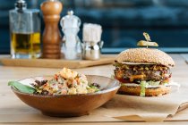 Великий гамбургер на папері з котлетиним сиром та овочами, подається з мискою барвистого салату в сучасному ресторані — стокове фото