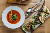 Вид сверху на красный суп с мясом и свежими травами на деревянном столе с кебабом и плоским хлебом в ресторане — стоковое фото