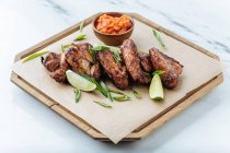 From above of grilled chicken wings on wooden board with slices of lime herbs and red sauce — Stock Photo