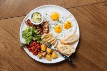 Vista dall'alto della colazione inglese servita con uova fritte e pancetta con pane tostato e verdure sul tavolo di legno — Foto stock