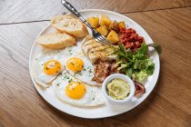 Vue du dessus du petit déjeuner anglais servi avec des œufs frits et du bacon avec des toasts et des légumes sur une table en bois — Photo de stock
